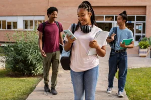 Três estudantes caminhando pelo pátio de uma instituição de ensino, com mochilas, cadernos e celular nas mãos, em frente a um prédio de tijolos.