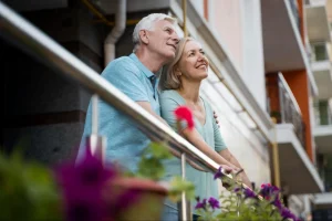 Casal idoso sorridente abraçado na varanda de apartamento, admirando a vista urbana com plantas roxas em vasos próximos, desfrutando de tranquilidade e conforto.