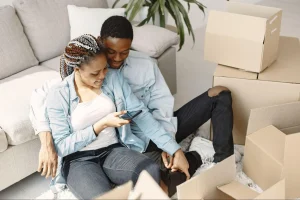 Casal jovem negro sorridente sentado no chão de novo apartamento durante mudança, cercado por caixas de papelão, plantas e sofá, interagindo com celular.