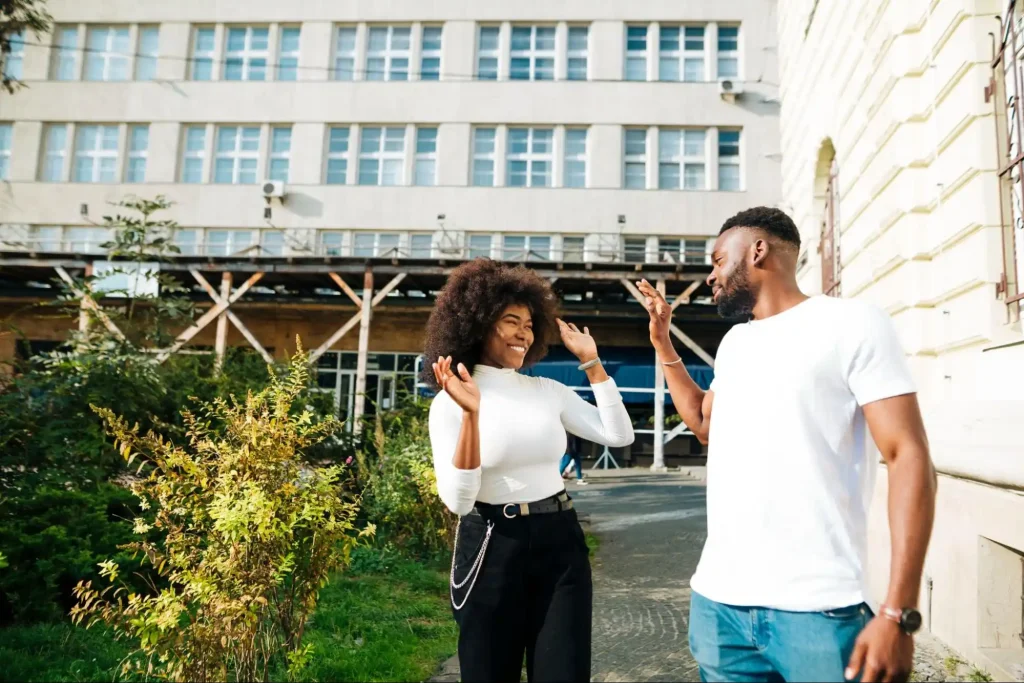 Casal jovem sorridente caminhando em área externa de condomínio com edifício ao fundo, cercados por vegetação, representando estilo de vida urbano moderno.