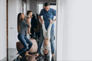 Família jovem com bebê e outro menino pequeno brincando em um apartamento moderno com piso de madeira, parede de tijolos aparentes e pufe colorido. O pai está levantando a criança de cabeça para baixo.