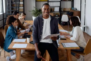 Homem sorridente no centro de equipe diversa em sala de reuniões moderna, segurando tablet, com colaboradores trabalhando em documentos e notebooks ao redor da mesa.