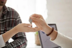 Mão feminina recebendo chaves de imóvel de mão masculina, em mesa de escritório com notebook aberto ao fundo, simbolizando a entrega de chaves no fechamento de contrato de aluguel.