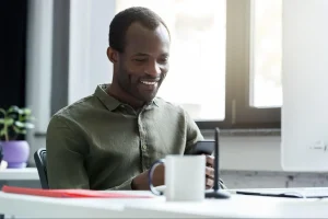 Homem negro sorridente em escritório luminoso segurando celular enquanto observa a tela, com xícara de café e planta roxa sobre a mesa ao lado.