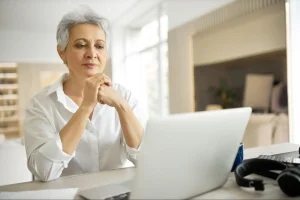 Mulher de meia-idade com cabelo curto grisalho sorrindo concentrada em laptop, com mãos sobre o teclado, em escritório moderno e iluminado por janela.