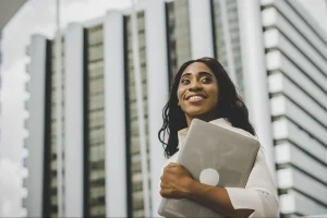 Mulher sorridente usando uma camisa branca e segurando laptop. Ela está em pé na frente de um edifício comercial moderno com fachada de vidro.