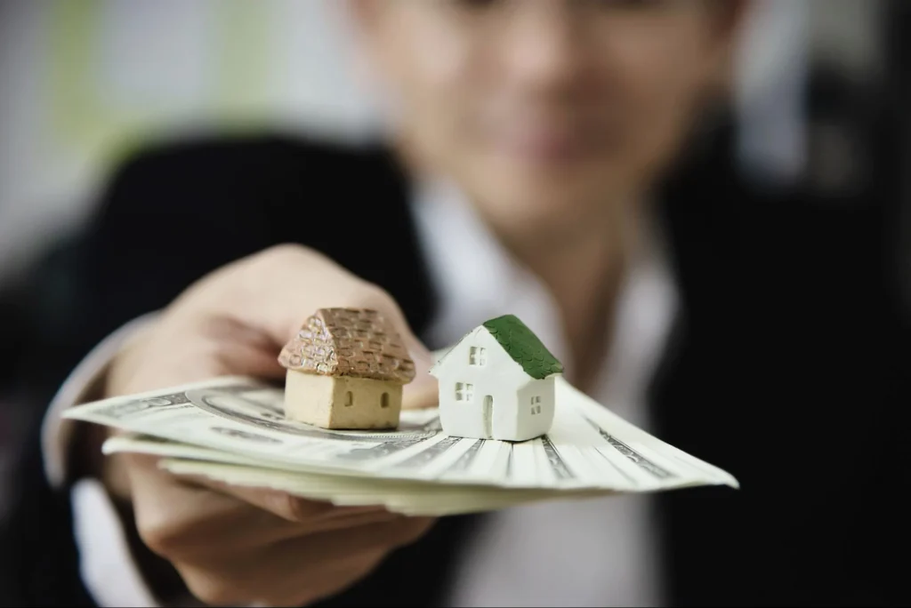 Pessoa segurando um maço de notas de dinheiro com duas miniaturas de casas apoiadas em cima, simbolizando a escolha entre diferentes investimentos imobiliários e financeiros.