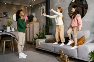 Três crianças de diferentes idades brincando com bolhas de sabão em sala de estar moderna com parede texturizada, sofá cinza, plantas verdes e brinquedos espalhados.