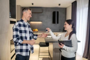Corretora sorridente entregando chaves de apartamento para homem de camisa xadrez em cozinha moderna com ilha central, frutas sobre bancada, cortinas claras e acabamento em cinza.