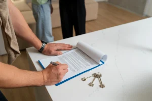 Homem assinando contrato imobiliário sobre bancada de quartzo branca com chaves ao lado, enquanto outra pessoa de calça preta observa ao fundo em ambiente residencial moderno.
