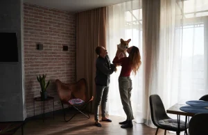 Homem e mulher sorridentes brincam com um cachorrinho Spitz em apartamento amplo de paredes de tijolo aparente, poltronas coloridas, mesa de jantar moderna, cortinas claras e luz natural suave entrando pelas janelas.