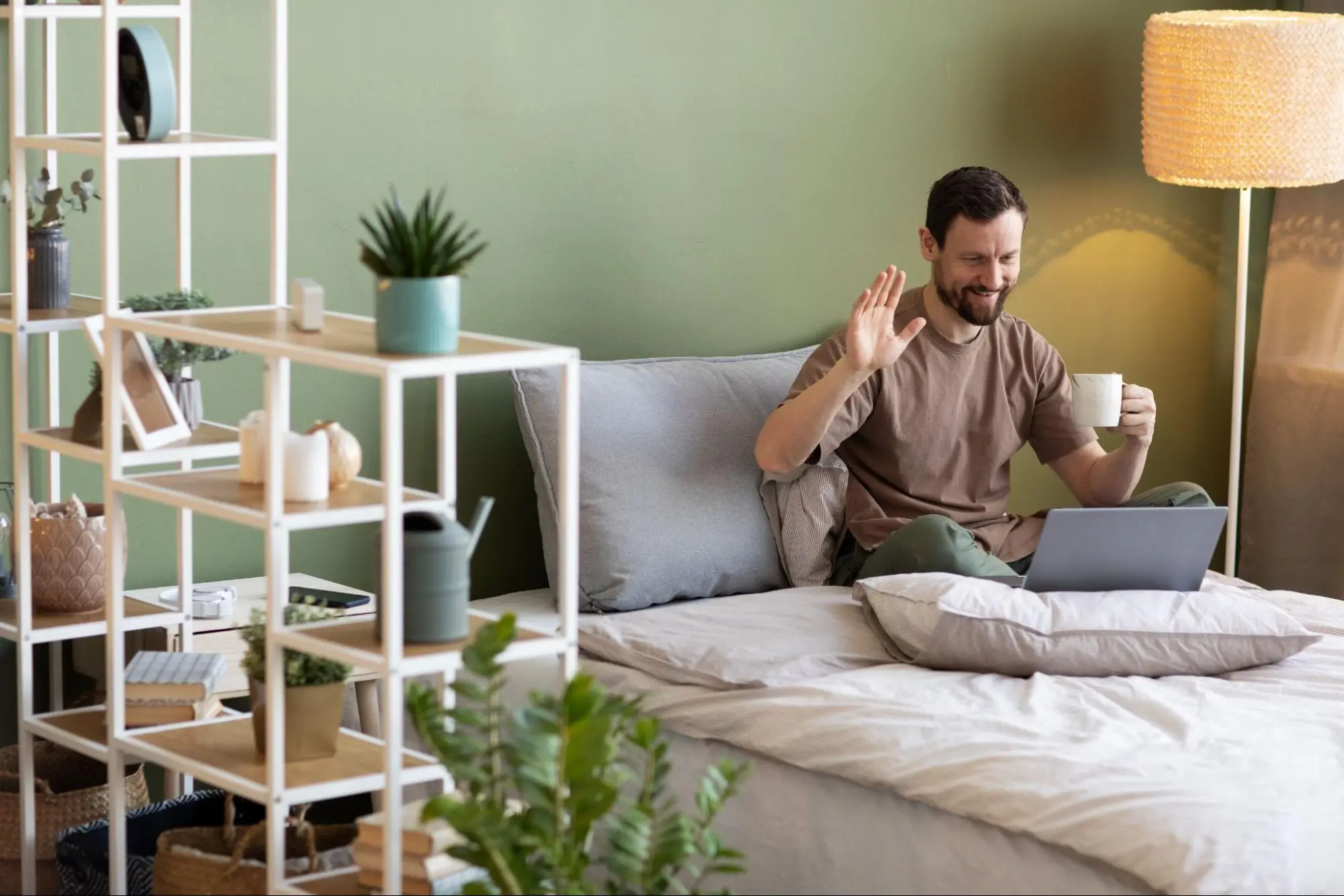 Homem sorridente acena para videoconferência no laptop da cama em quarto compacto moderno de parede verde, prateleira branca com plantas, vasos, relógio, livros, abajur trançado e luz suave