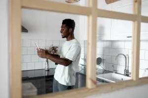 Homem sorridente de camiseta na cozinha compacta moderna com azulejos brancos, bancada preta, pia branca, cooktop e armários brancos.