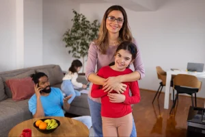 Mãe sorridente de óculos abraçando filha de blusa vermelha em sala de estar ampla, pai ao fundo no sofá com celular e frutas na mesa, outra menina brincando, ambiente moderno com planta, poltronas e piso de madeira.