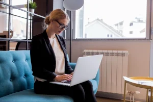 Mulher de blazer preto usa laptop sentada em sofá azul, em ambiente iluminado, com janela ao fundo.