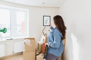 Mulher de camisa azul organiza decoração em apartamento compacto. Ela observa um quadro de planta verde, cercada de caixas de mudança, janelas amplas com luz natural e radiadores ao lado do piso de madeira.