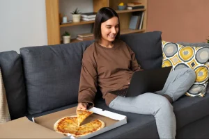 Mulher sorridente de cabelo curto castanho come fatia de pizza de caixa aberta no sofá cinza com almofadas geométricas multicoloridas, laptop no colo, estante de madeira ao fundo com livros em apartamento compacto moderno.