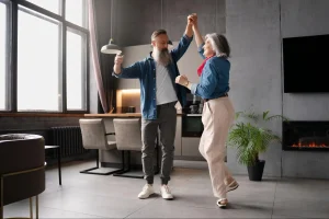 Um casal de idosos dança sorridente no meio de uma sala de estar moderna e ampla. O ambiente possui decoração em tons de cinza, grandes janelas que iluminam o espaço, uma planta decorativa e uma lareira elétrica ao fundo, transmitindo uma sensação de conforto e felicidade.