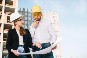 Uma mulher com capacete branco e blazer preto e um homem com capacete amarelo e camisa branca seguram e analisam atentamente uma planta arquitetônica. Eles estão em um canteiro de obras, com a estrutura de um prédio em construção ao fundo sob um céu claro.