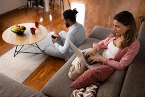 Vista em ângulo alto de casal relaxando na sala ampla do apartamento. A mulher está de camisa rosa e trabalha no laptop no sofá cinza com almofadas coloridas, enquanto o homem está sentado no chão com o celular nas mãos.