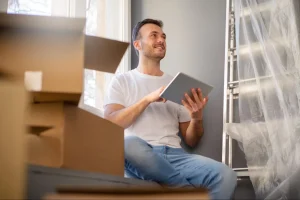 Homem sorridente, de camiseta branca e calça jeans, sentado em um apartamento novo durante a mudança. Ele segura um tablet e olha para o lado com otimismo, cercado por caixas de papelão e uma escada de alumínio coberta por plástico.