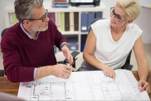 Um homem de cabelos grisalhos e suéter bordô e uma mulher loira de blusa branca, ambos de óculos, estão sentados à mesa analisando uma planta baixa de arquitetura.