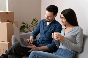 Um jovem casal sorri enquanto usa um notebook juntos, sentados em um sofá cinza. O homem veste camisa jeans azul e a mulher, de blusa cinza, segura uma caneca branca. Ao fundo, caixas de papelão empilhadas indicam uma mudança recente, transmitindo a sensação de um novo começo residencial.