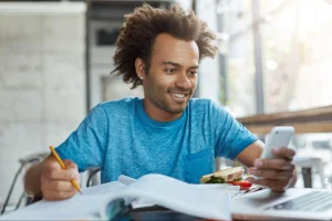 Homem sorri ao olhar o smartphone enquanto faz anotações em um caderno. Ele está em um local iluminado, com um sanduíche e parte de um notebook sobre a mesa.
