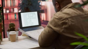 Homem visto de costas analisa a planta baixa detalhada de um apartamento na tela de um notebook aberto sobre a mesa. Ao lado do computador, há plantas impressas em papel, um copo de café descartável e um pequeno cacto em um escritório iluminado.