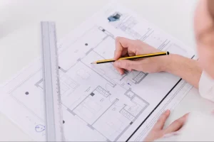 Imagem em plano detalhado com foco nas mãos de uma pessoa analisando a planta de um imóvel sobre uma mesa branca. Ela segura um lápis amarelo e preto apontando para o desenho técnico, enquanto uma régua de metal está posicionada ao lado do papel.