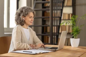 Mulher de cabelos grisalhos sorri levemente enquanto usa um notebook em uma mesa de madeira. Ela usa fones de ouvido sem fio, casaco bege claro e tem óculos de grau apoiados sobre papéis na mesa. Ao fundo, uma estante de livros e um vaso de planta decoram o ambiente iluminado.