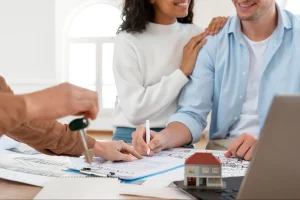 Casal sorridente assina um contrato de compra de imóvel sobre uma mesa com plantas arquitetônicas e uma miniatura de casa. O homem segura uma caneta branca enquanto uma terceira pessoa, à esquerda, aponta para o documento e segura um molho de chaves.